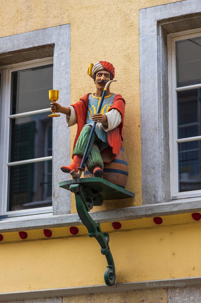 A carved wooden man in Turkish attire offering a meter-long pipe and a golden goblet from a platform on a wall in Solothurn, Switzerland.