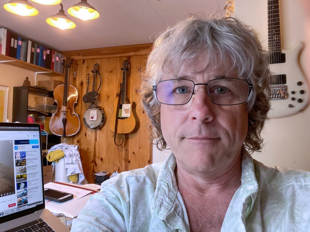 John's face is friendly; he has squarish glasses, grey hair and is clean shaven. In the background are a cello, banjo and guitar hanging on the wall, and a computer open on his desk.