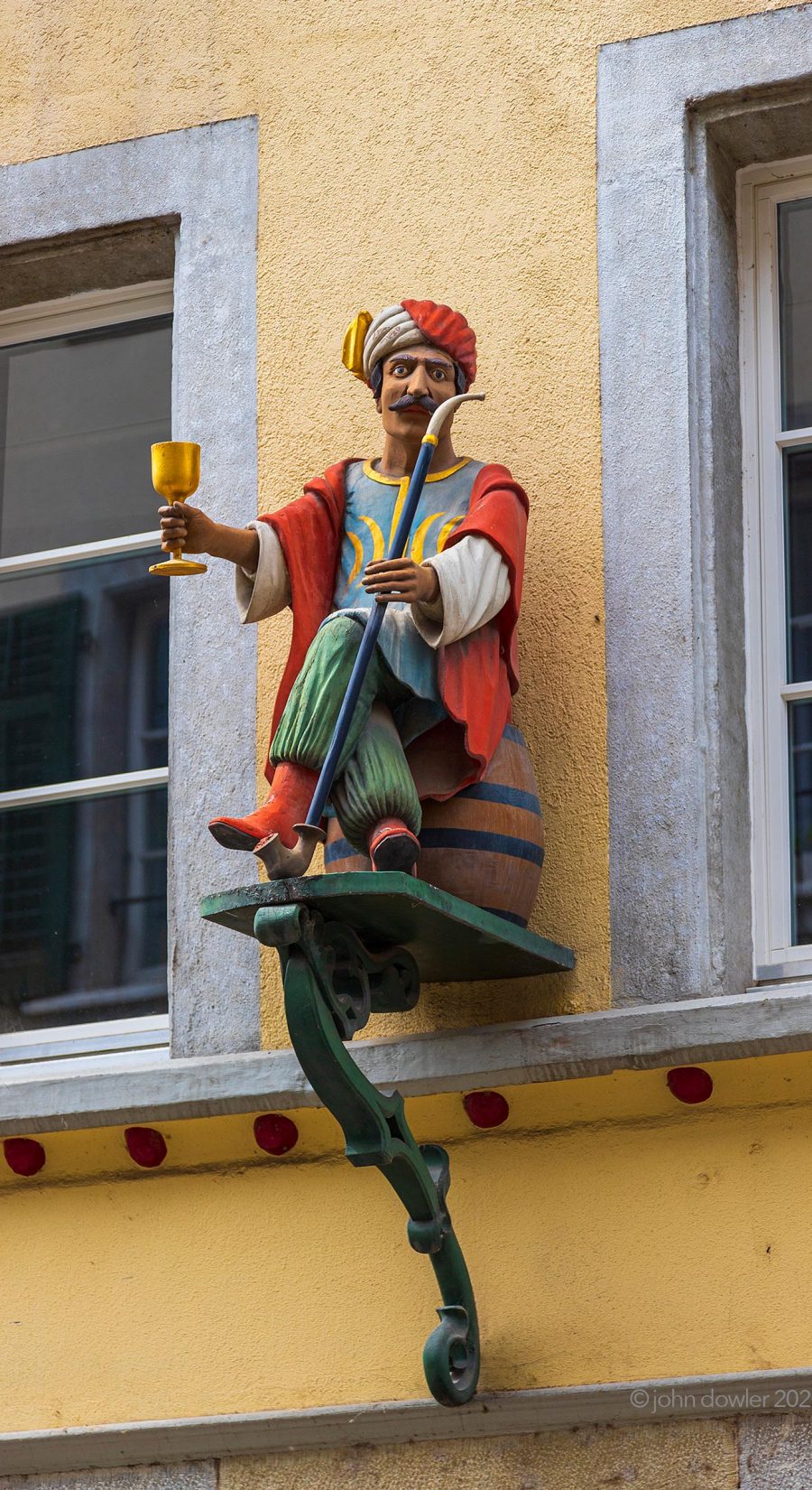 A carved wooden man in Turkish attire offering a meter-long pipe and a golden goblet from a platform on a wall in Solothurn, Switzerland.